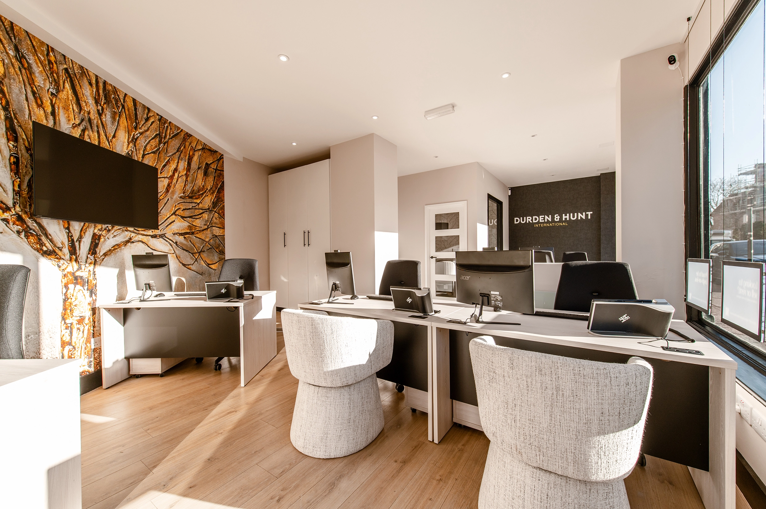 Modern office interior with three desks, beige chairs, computer monitors, and a dark accent wall with "DURDEN & HUNT INTERNATIONAL" signage. Gold-patterned wallpaper and TV screen on left wall.