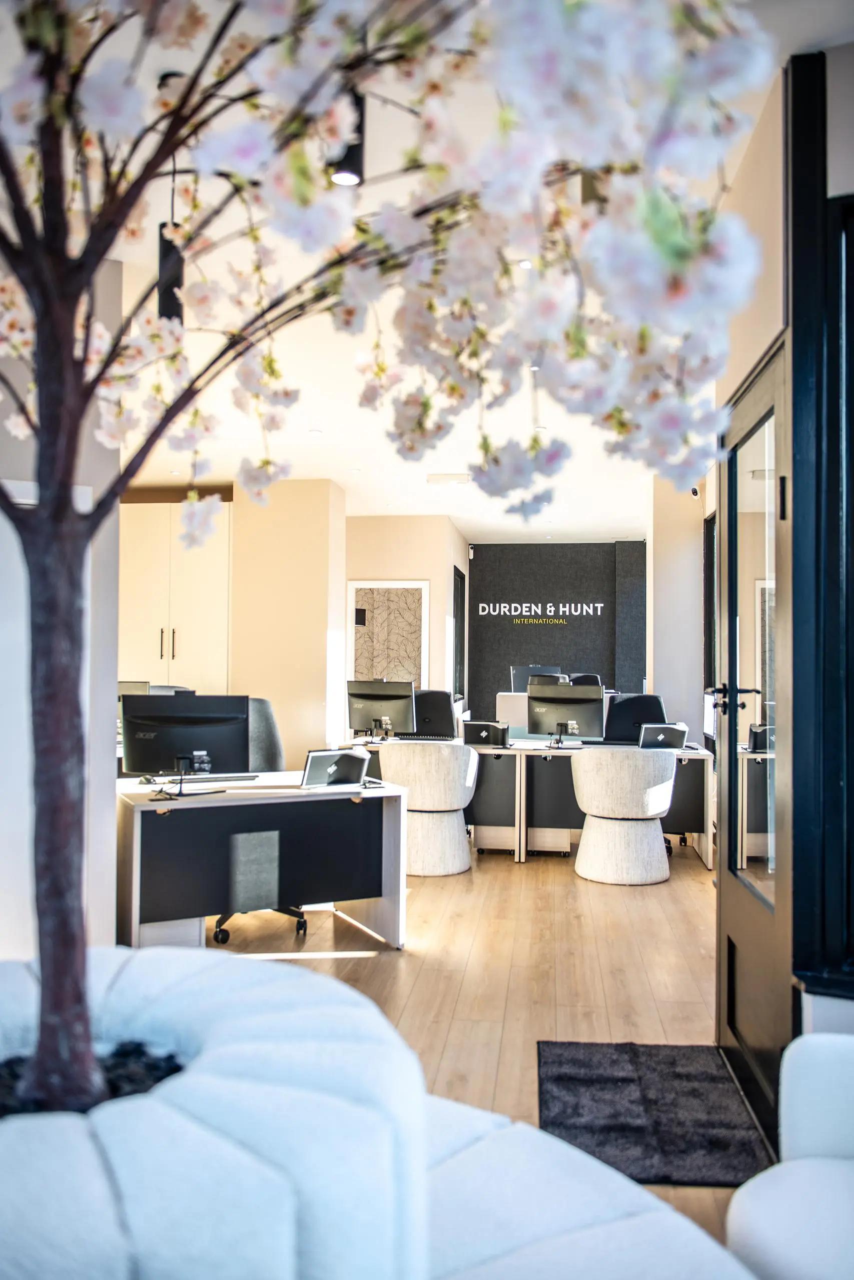 Modern office interior with "Durden & Hunt International" signage on dark wall, desks with computer monitors, cream chairs, wooden flooring, and flowering tree branches overhead