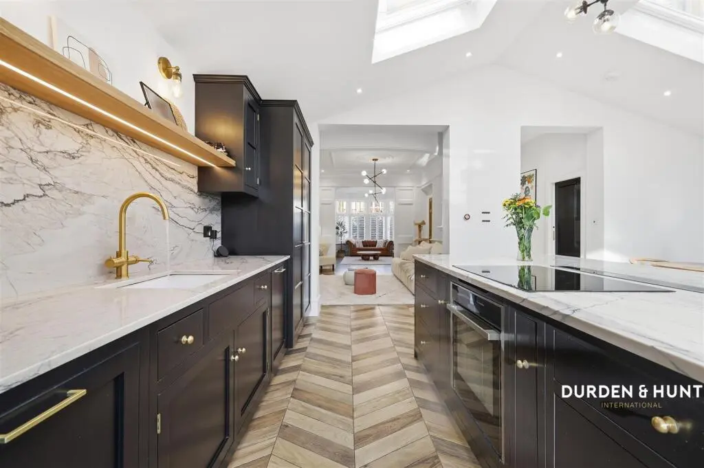 Modern kitchen with dark cabinetry, marble worktops, brass fixtures, and herringbone wood flooring, opening into a bright living space with skylight
