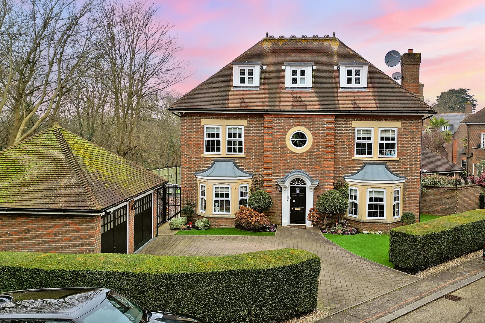 Red brick detached house with mansard roof, three dormer windows, bay windows, black front door, and brick driveway with garage to the left