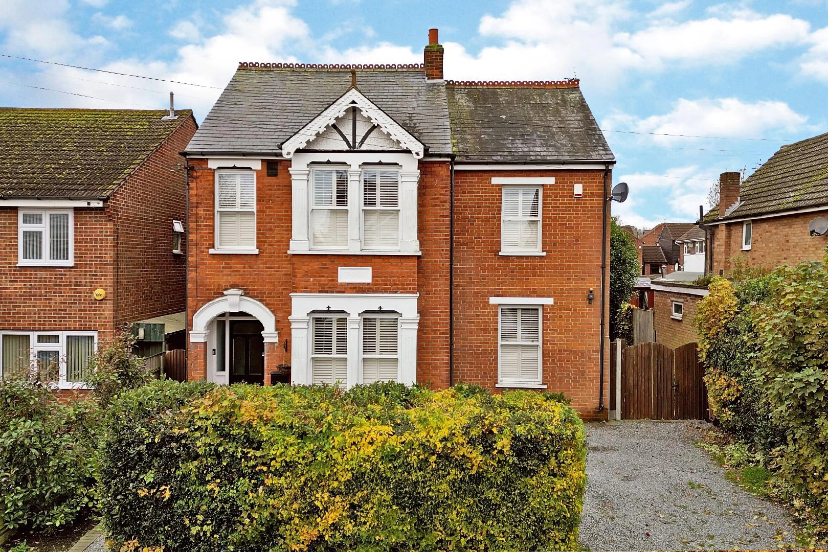 Red brick detached house with white-framed windows, arched entrance, slate roof and brick chimney, set back behind hedges