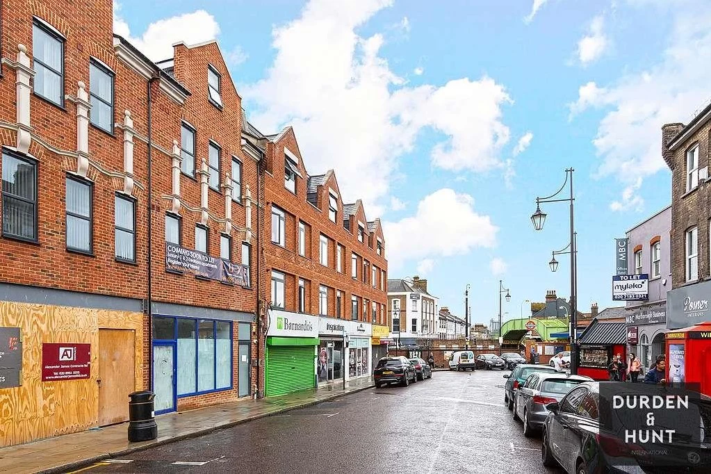 Street view of brick buildings with shops at ground level, including a green storefront and boarded-up windows, parked cars, and street lamps under blue sky