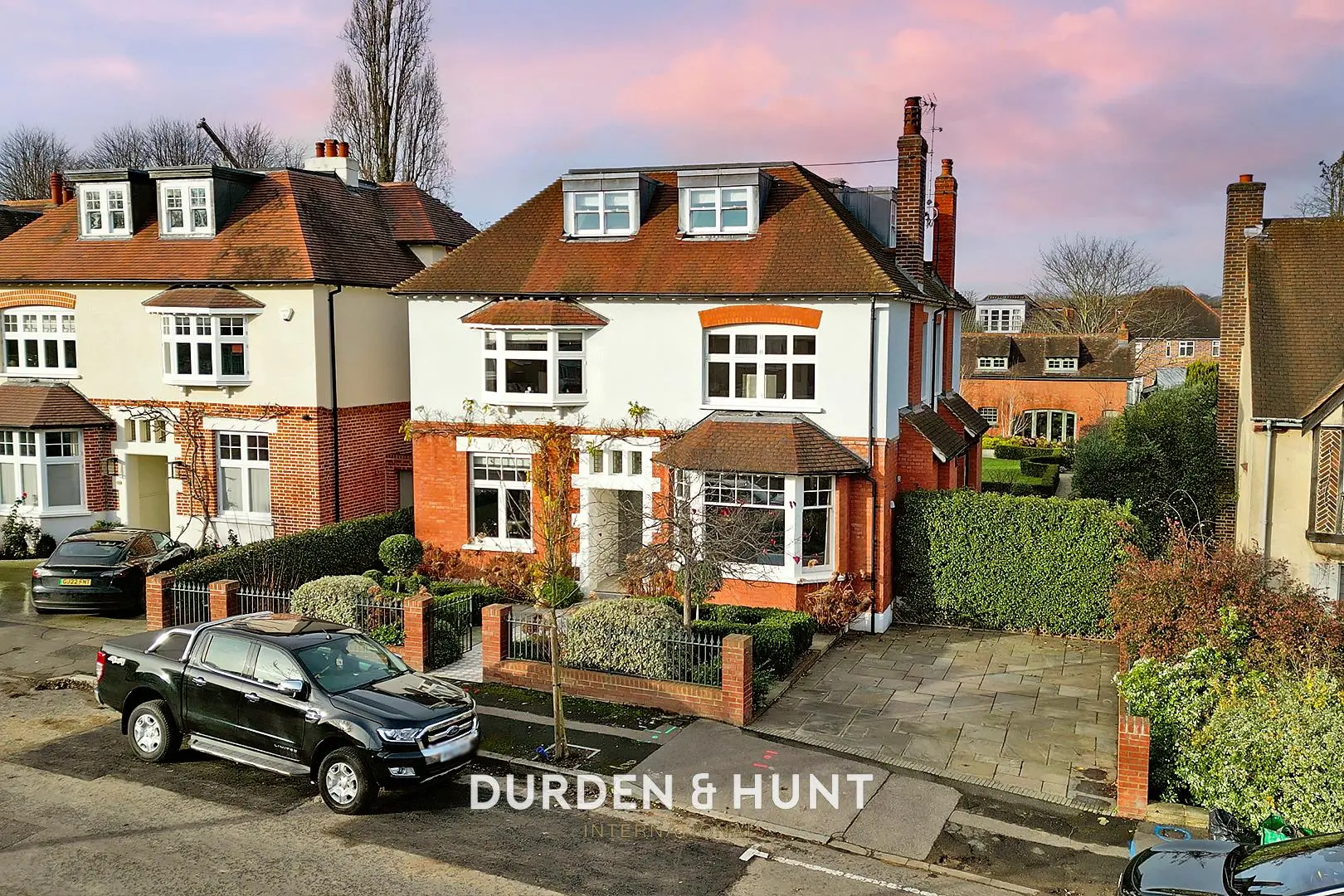 Detached house with red brick and cream rendered walls, pitched tiled roof with dormer windows, and brick boundary wall. Black pickup truck parked in front. "Durden & Hunt" branding visible.