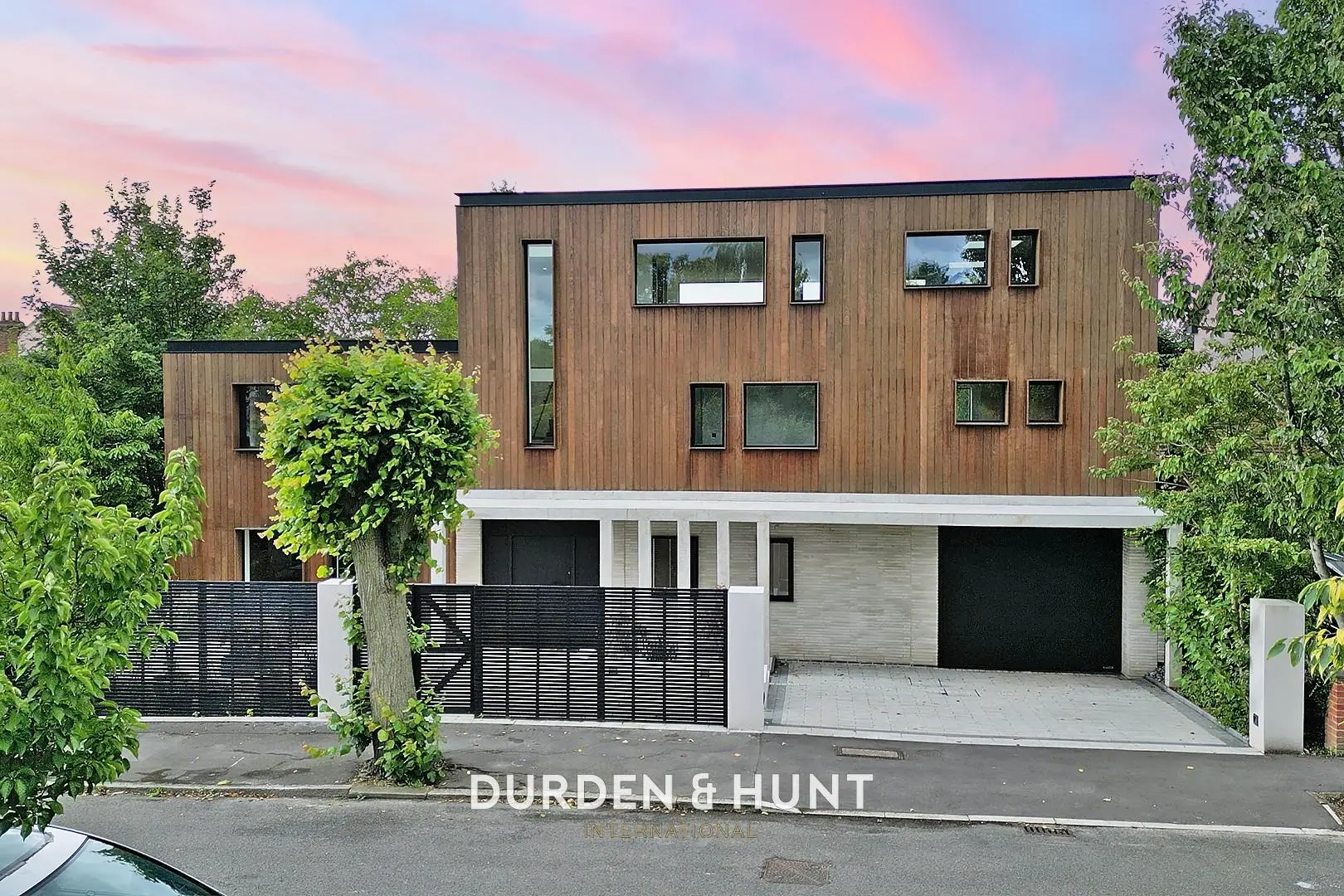 Modern two-storey house with brown timber cladding, white ground floor, black garage doors, black metal gates, and surrounding greenery