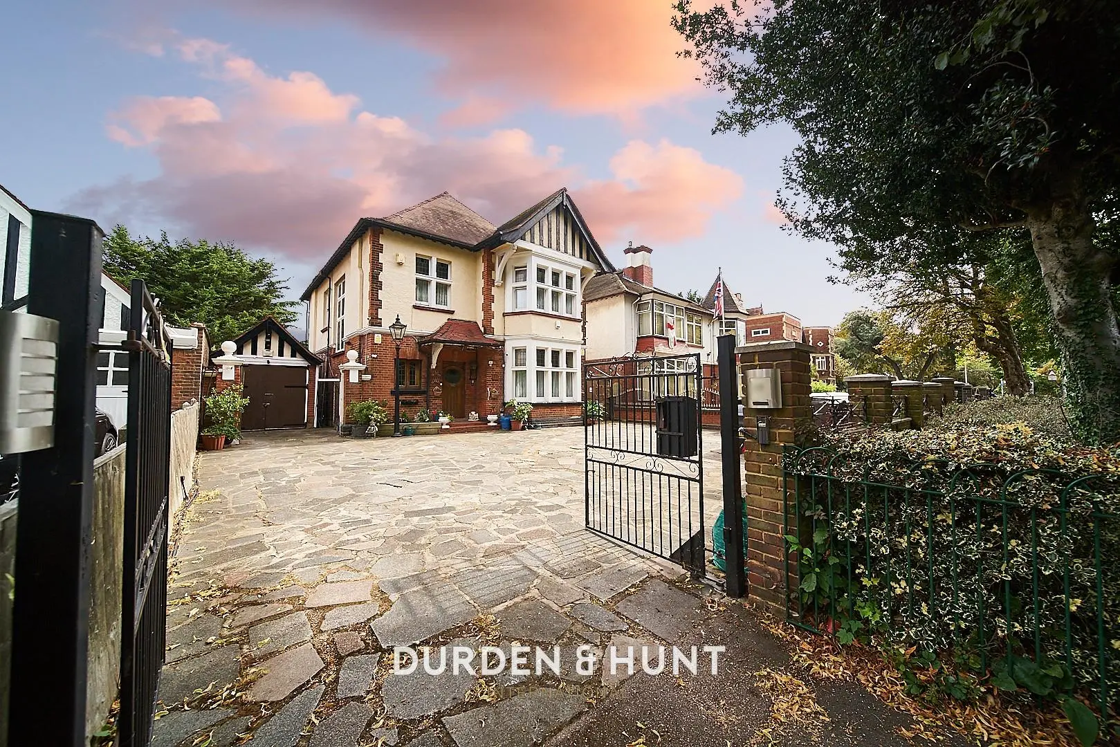 Cream and brown detached house with black metal gates and driveway, surrounded by trees and neighbouring properties
