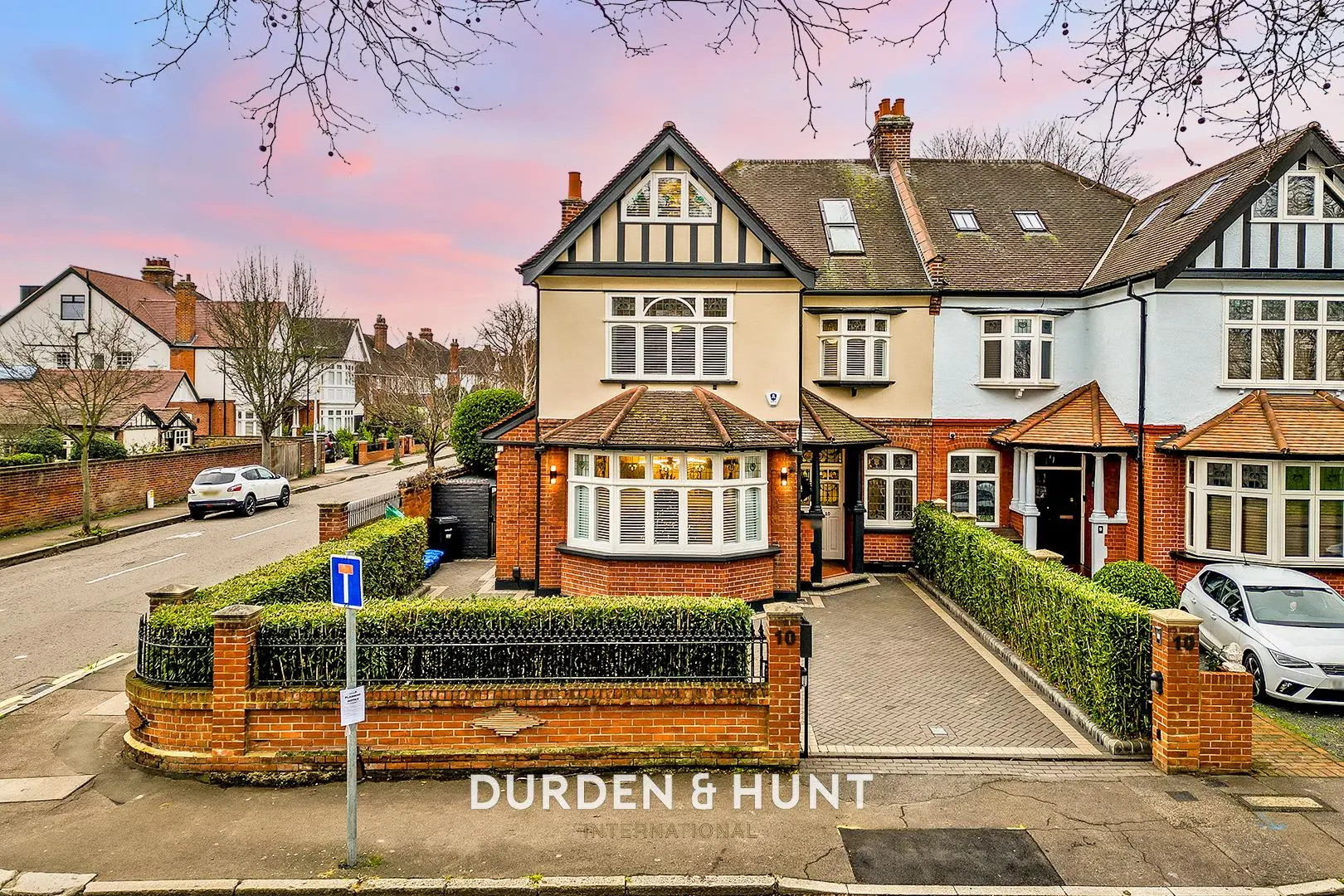 Cream and black half-timbered house with brick extension and driveway, neighbouring period properties visible on residential street at dusk