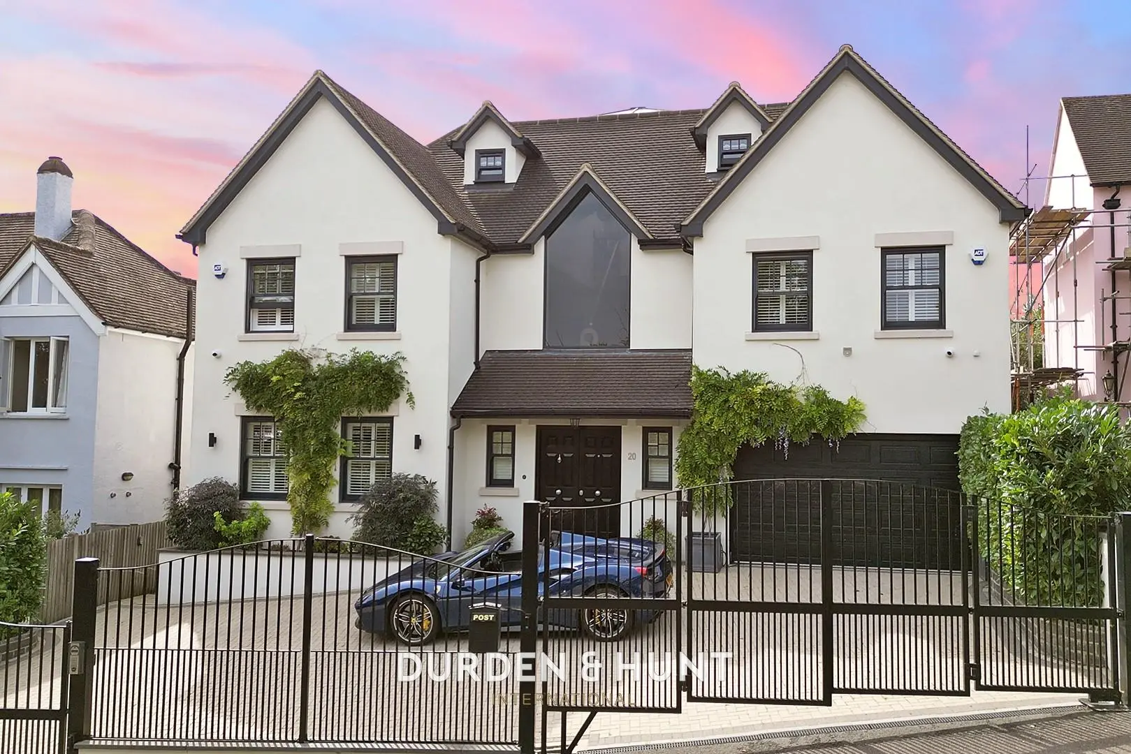 Modern white house with multiple gables, dark roof, black metal gates and driveway, parked car visible, pink sunset sky
