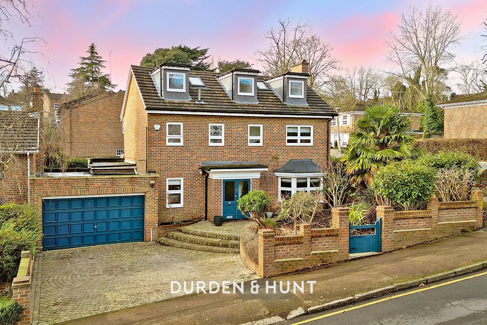 Brick house with blue garage door and entrance door, three dormer windows on roof, brick boundary wall with blue gate, driveway and front garden with plants