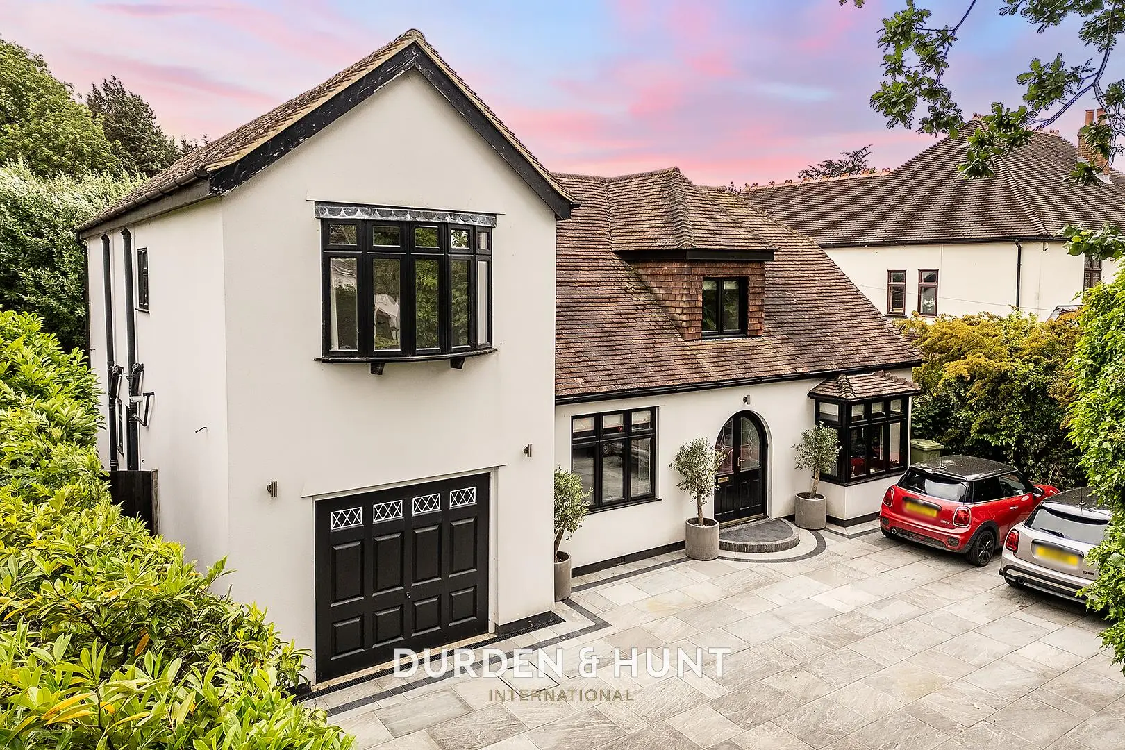 Modern white-rendered house with black windows and garage, tiled driveway, parked red car, and manicured hedges