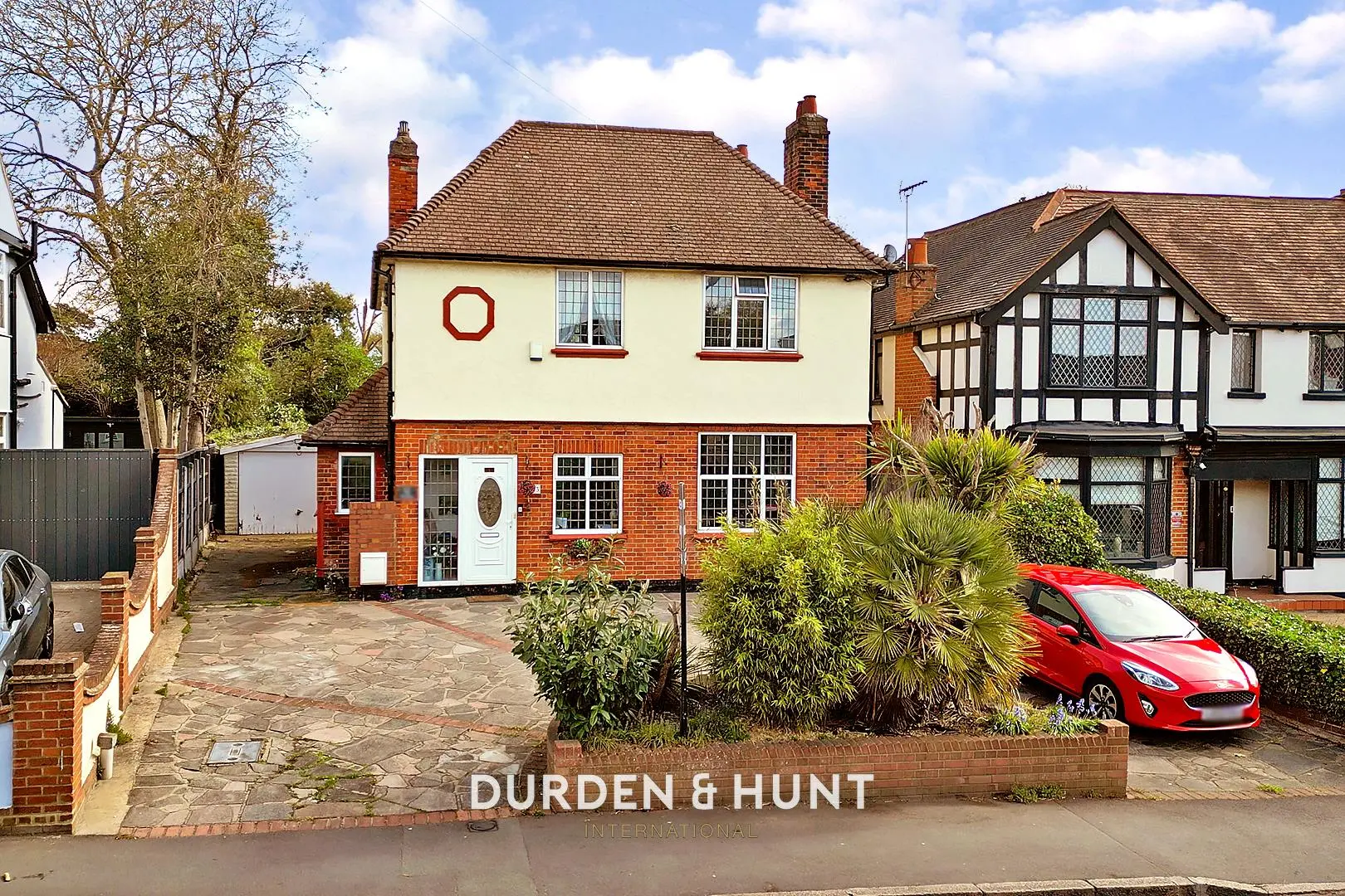 Two-storey detached house with cream-coloured upper floor and red brick lower floor, featuring a red octagon decoration, parked red car in front, and neighbouring Tudor-style property. Durden & Hunt branding visible.