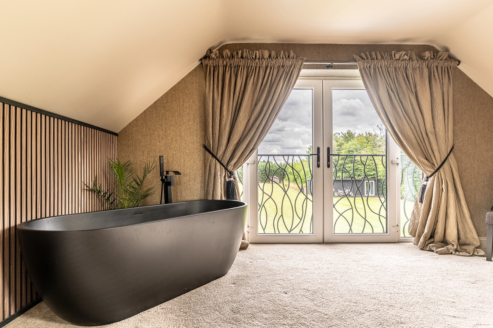 Attic bathroom with freestanding black bath, beige curtains, French doors opening to garden, and sloped ceiling