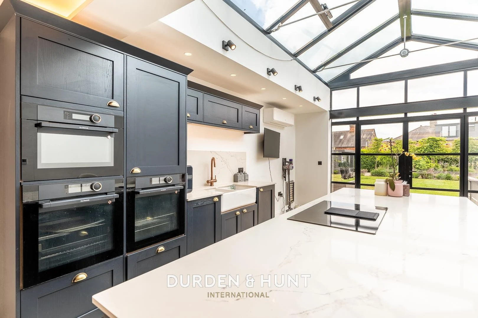 Modern kitchen with dark grey cabinetry, white marble countertops, integrated appliances, skylight roof, and glass doors opening onto a garden