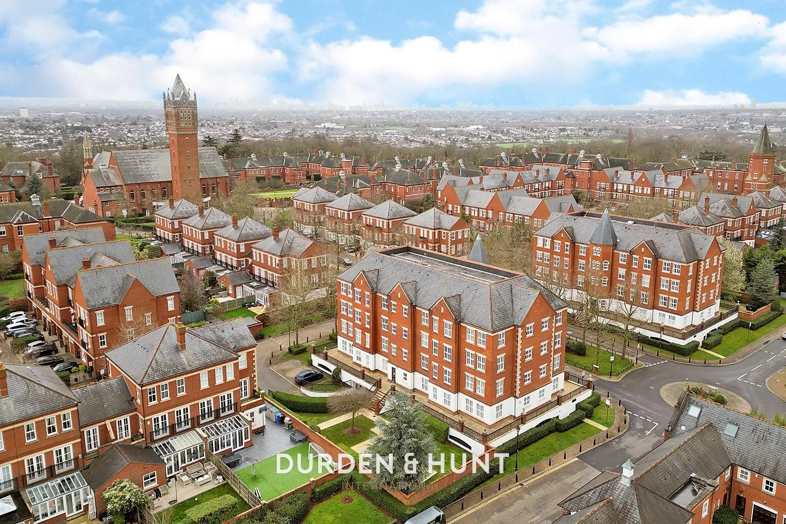 Aerial view of a large residential complex with red-brick Victorian buildings, manicured gardens, and a clock tower in the background