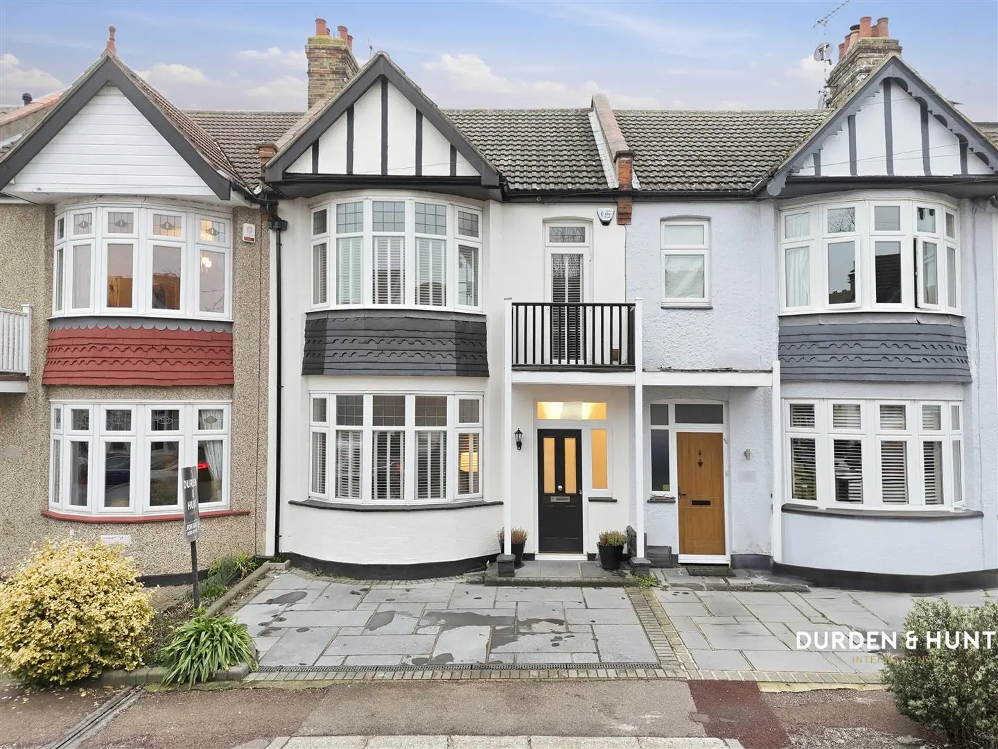 Row of three terraced houses with white and cream rendered walls, black and white timber-framed gables, tiled roofs, and bay windows. Centre house has black door with yellow surround; left house has red garage door; right house has brown door. Paved front forecourt with potted plants.