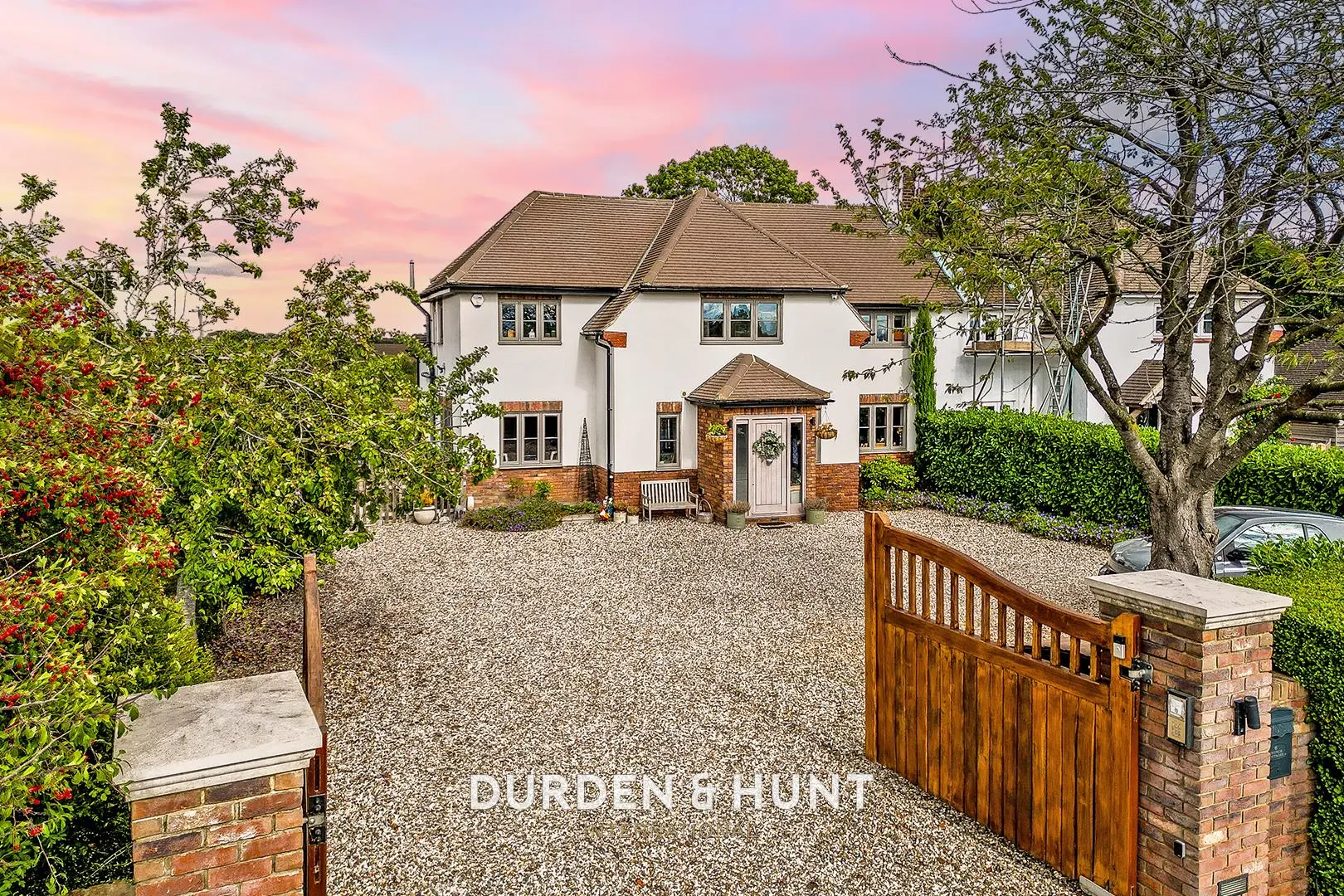 White two-storey house with brown tiled roof, brick entrance porch, gravel driveway, wooden gate, and surrounding trees and shrubs under pink sunset sky