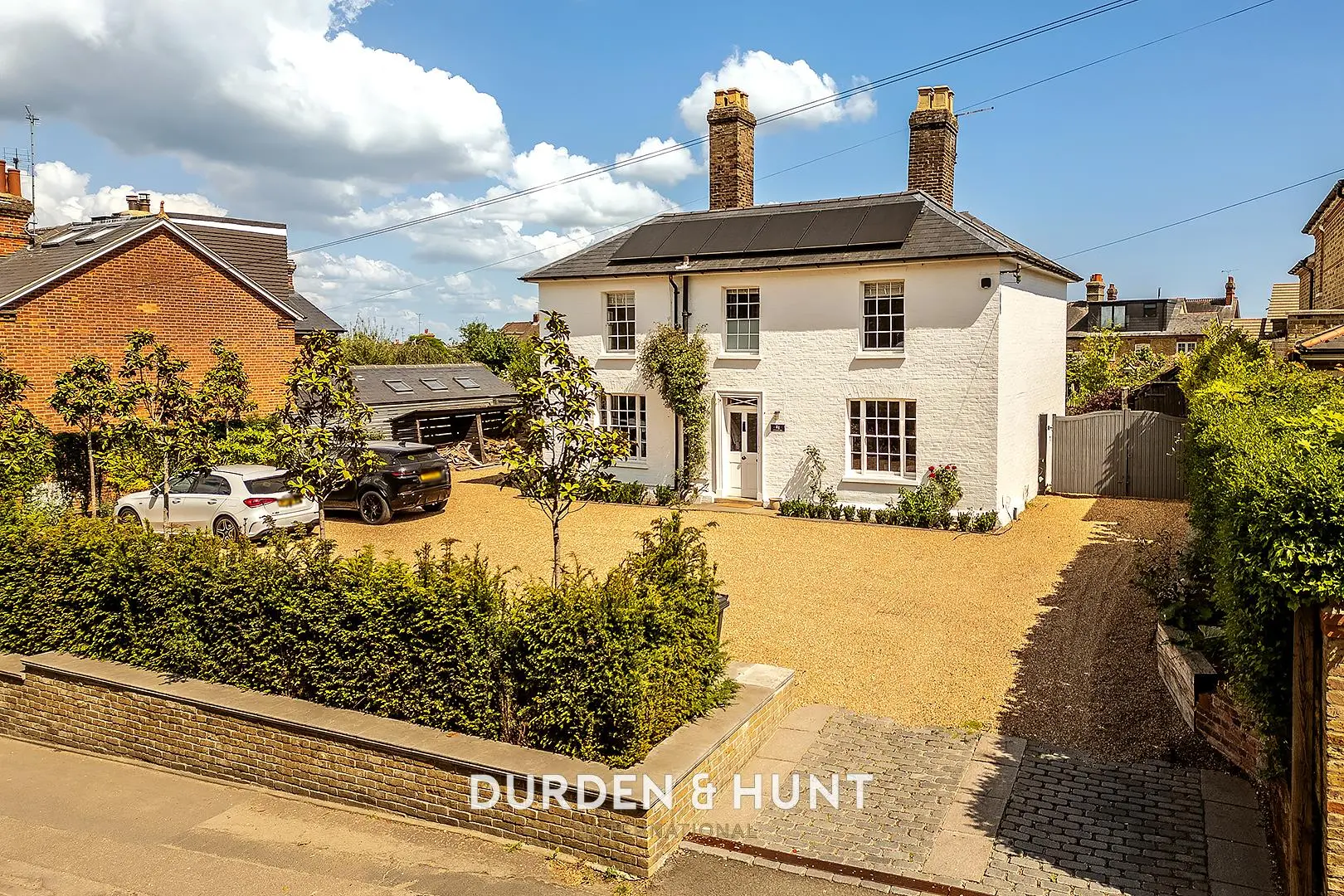 White rendered two-storey house with brick chimneys, gravel driveway with two parked cars, hedging and mature trees, neighbouring brick properties visible
