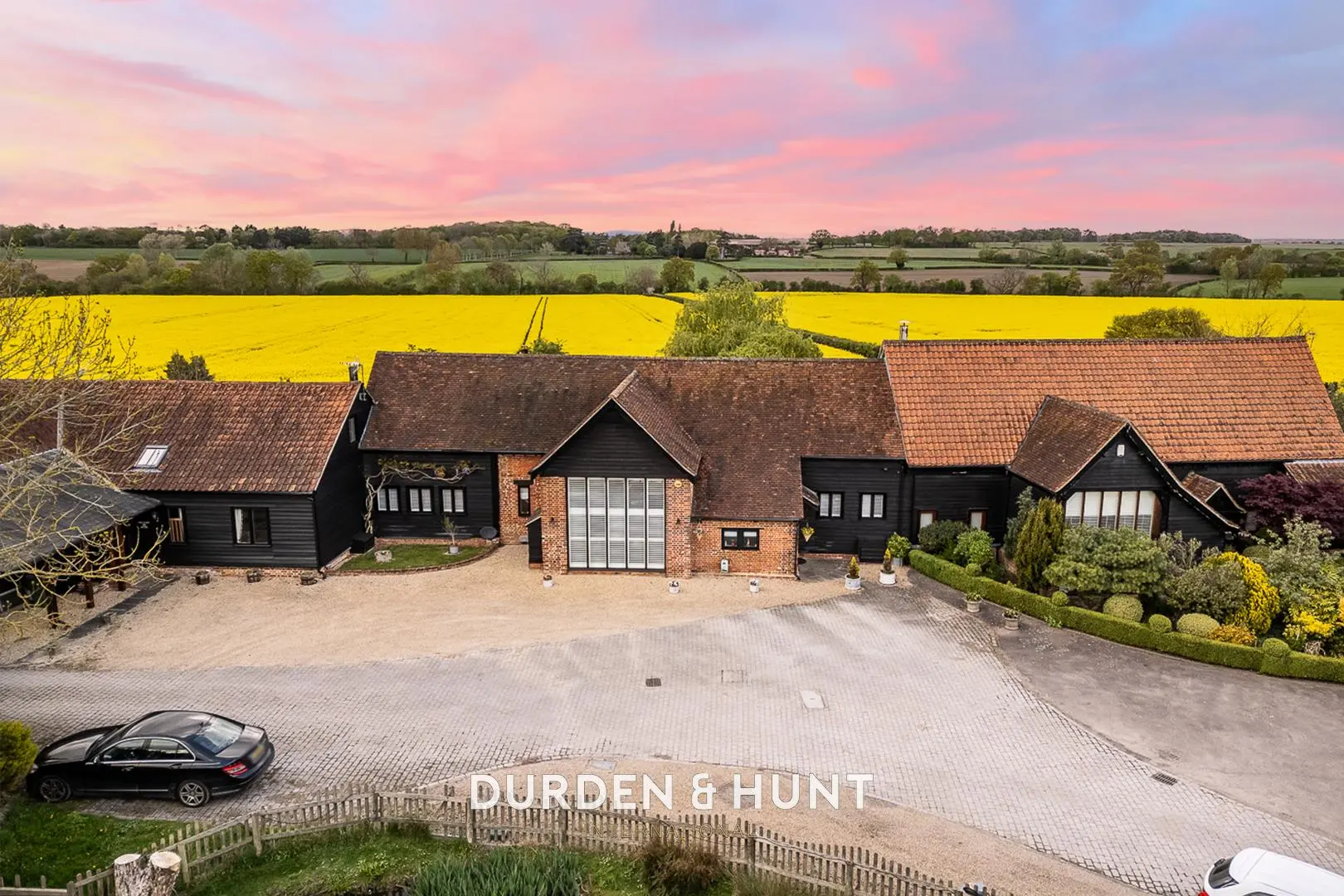 Aerial view of a converted barn complex with brick and black timber buildings, paved courtyard, and yellow rapeseed fields beyond. Durden & Hunt branding visible.