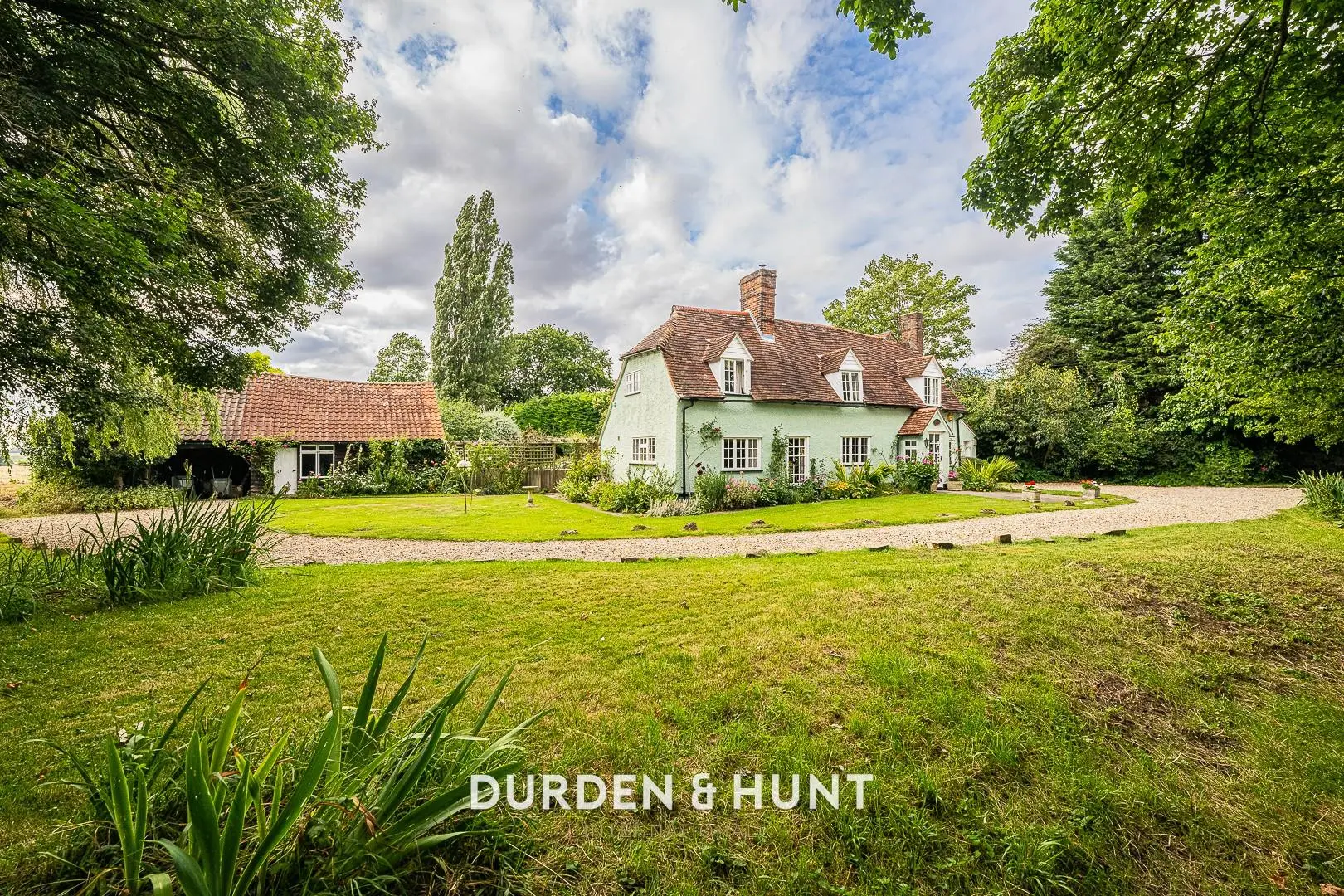 Cream-coloured country house with terracotta roof and ivy-covered walls, set in a large garden with manicured lawn and gravel driveway, surrounded by mature trees