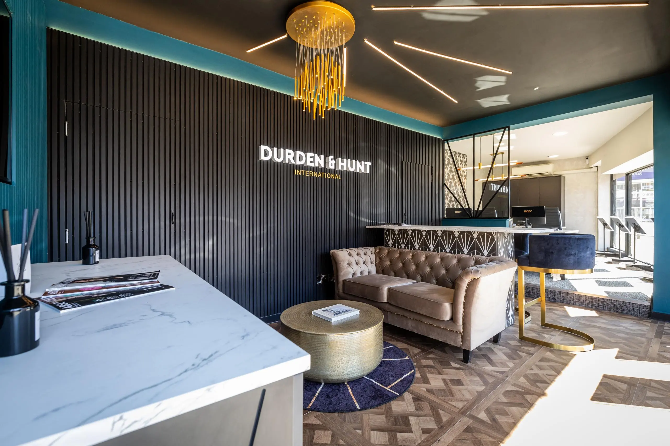 Durden & Hunt International estate agency office interior with marble reception desk, taupe sofa, gold pendant light, and teal accent walls