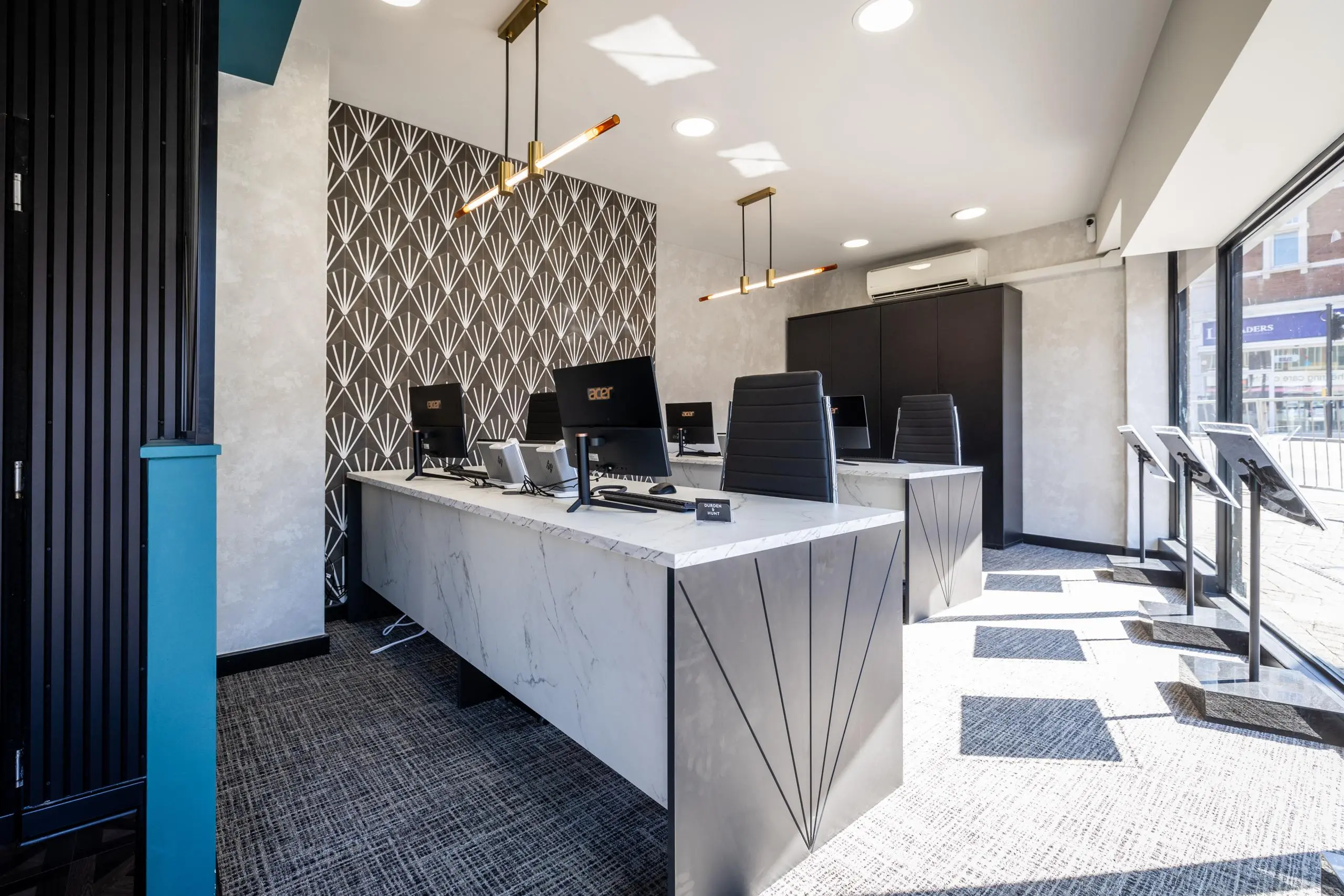 Modern office reception area with marble counter, computer monitors, geometric patterned wallpaper, pendant lights, and street-facing windows