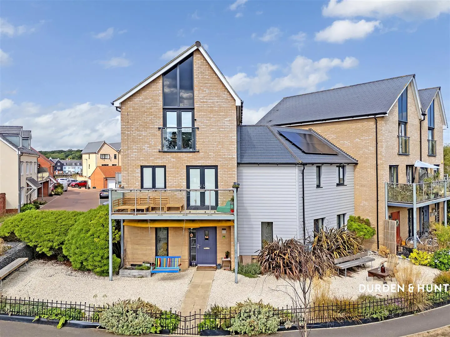 Modern brick house with gabled roof, large windows, decked terrace, and white garage extension. Solar panels visible on roof. Landscaped front garden with shrubs and black metal fencing.