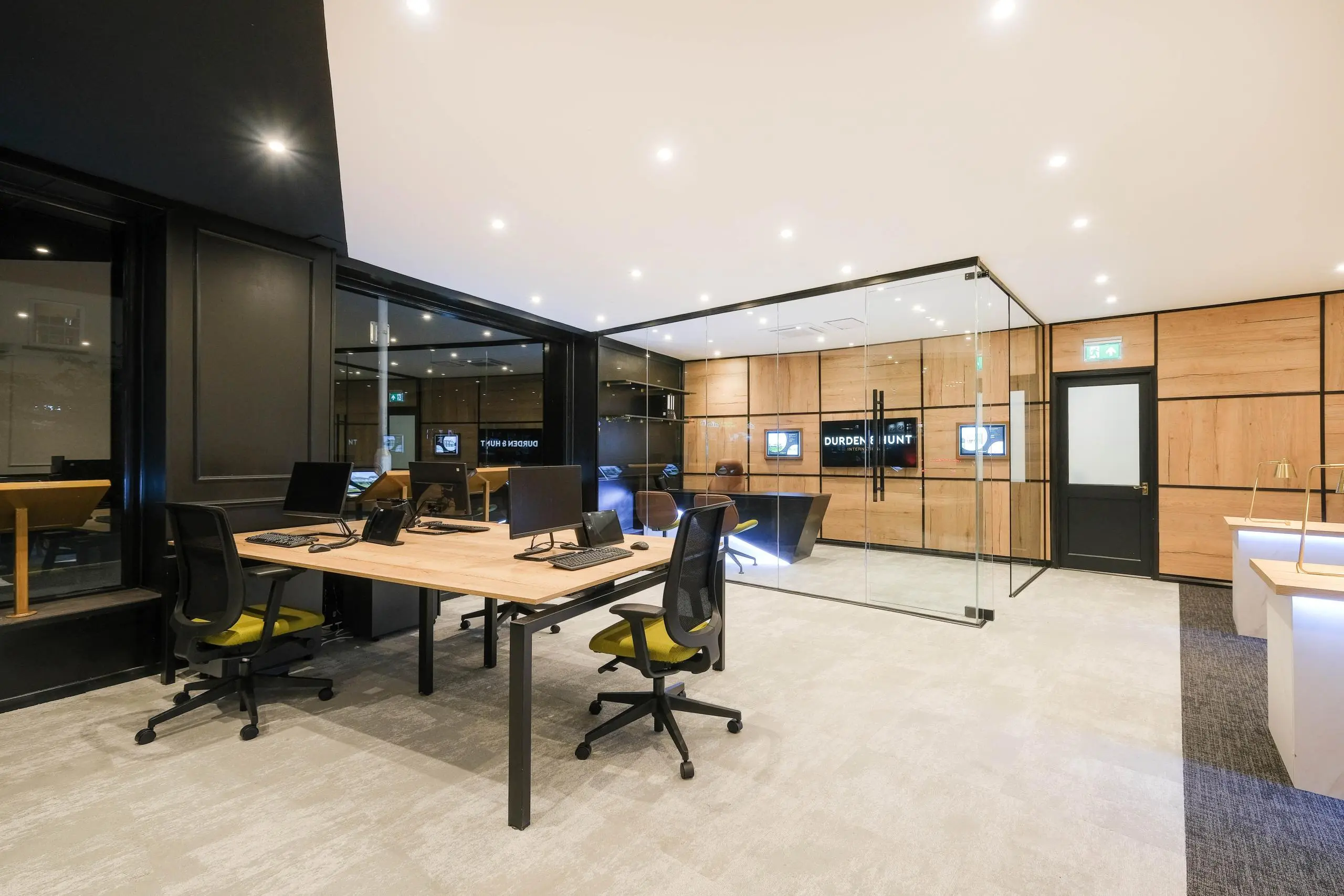Modern office interior with wooden and glass partitions, desk with computers and black chairs with yellow accents, recessed ceiling lights