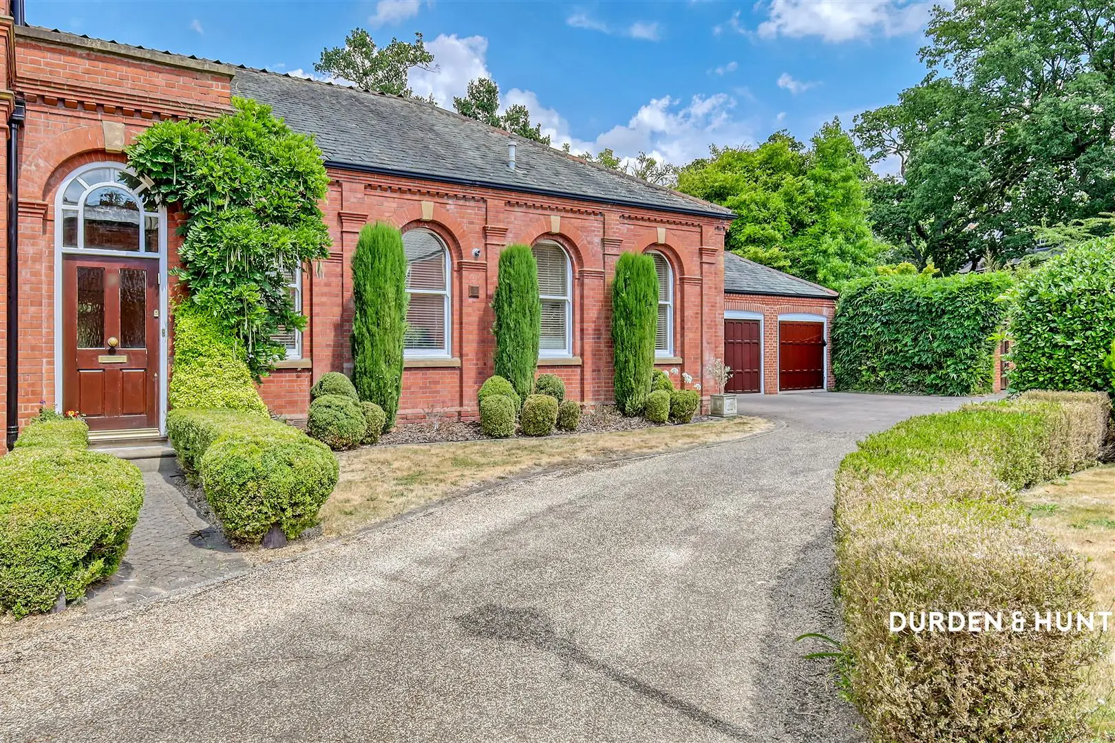 Red brick building with arched windows, ivy-covered walls, manicured topiary and cypress plants, driveway, and detached garage