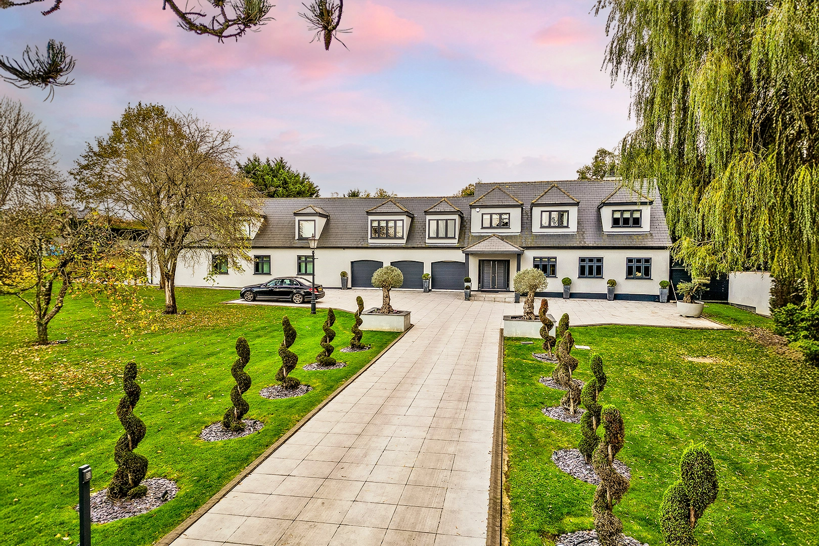 Modern two-storey house with mansard roof, multiple dormer windows, black garage doors, and paved driveway flanked by manicured topiary and lawns