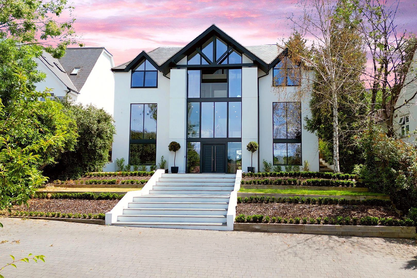 Modern white house with black-framed gable windows, dark front door, and wide stone steps leading to entrance, surrounded by landscaped gardens and trees