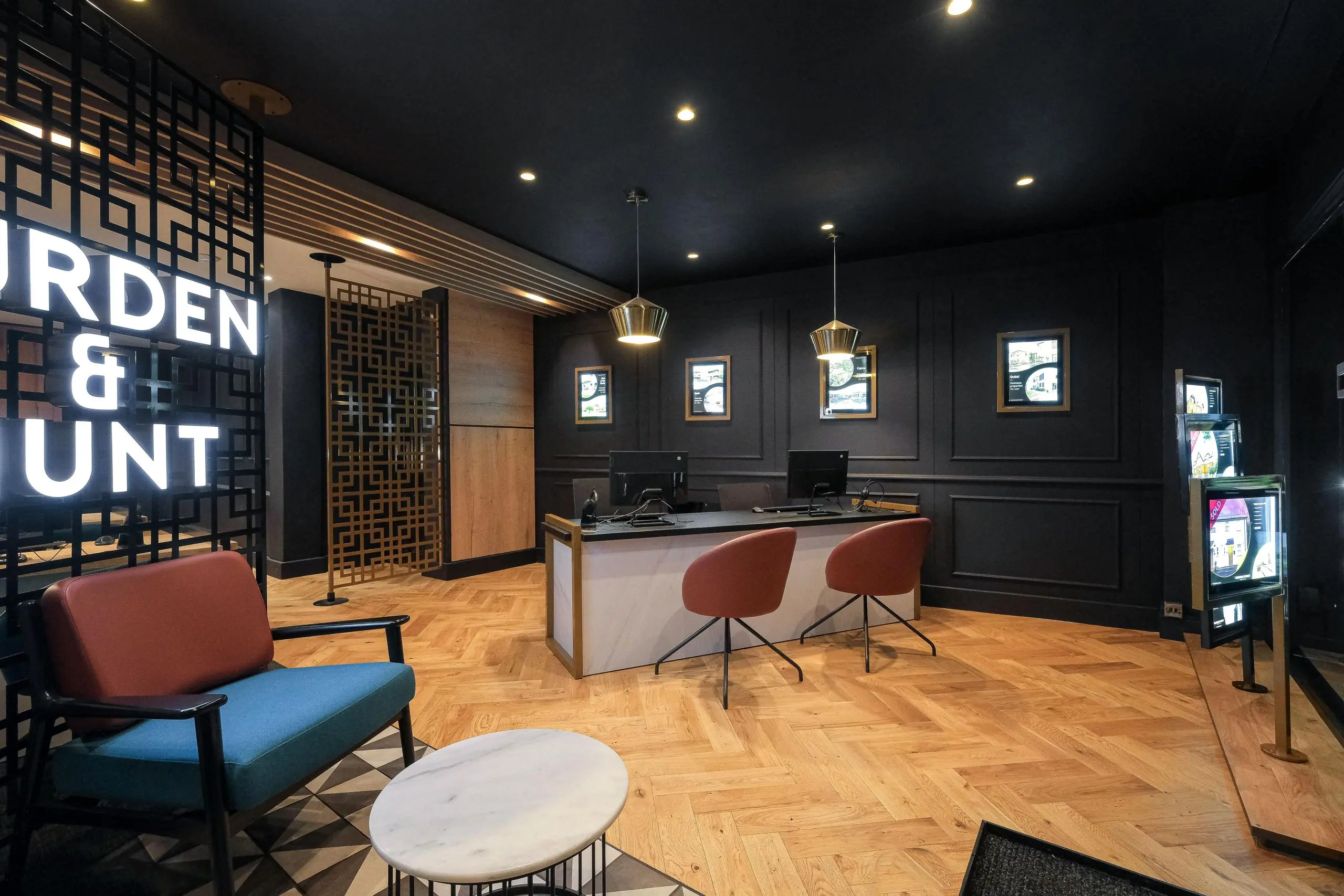 Modern office reception area with dark walls, wooden herringbone flooring, illuminated signage reading "RDEN & UNT", desk with red chairs, and seating area with blue and red upholstered furniture