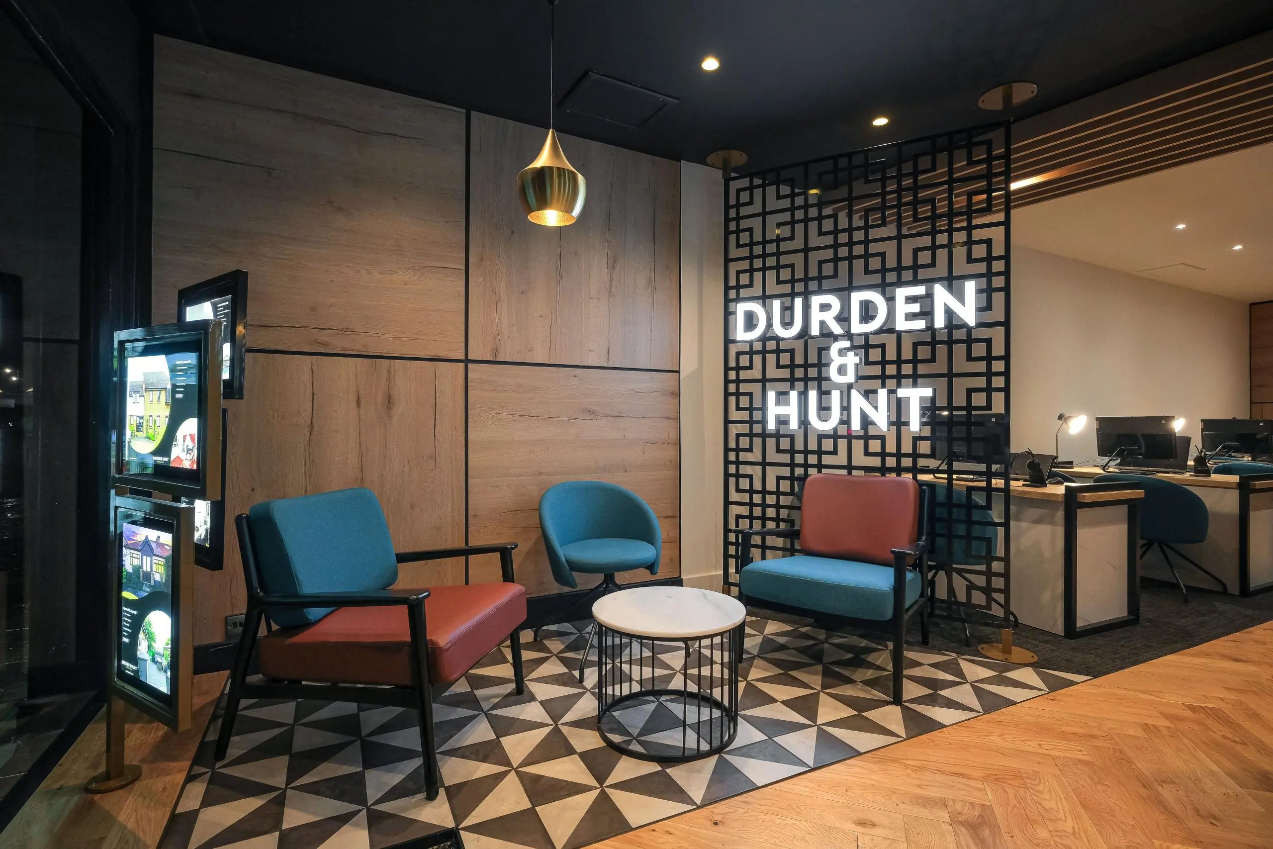 Modern office reception area with "Durden & Hunt" signage, blue and red mid-century chairs, geometric patterned floor, and wooden wall panelling