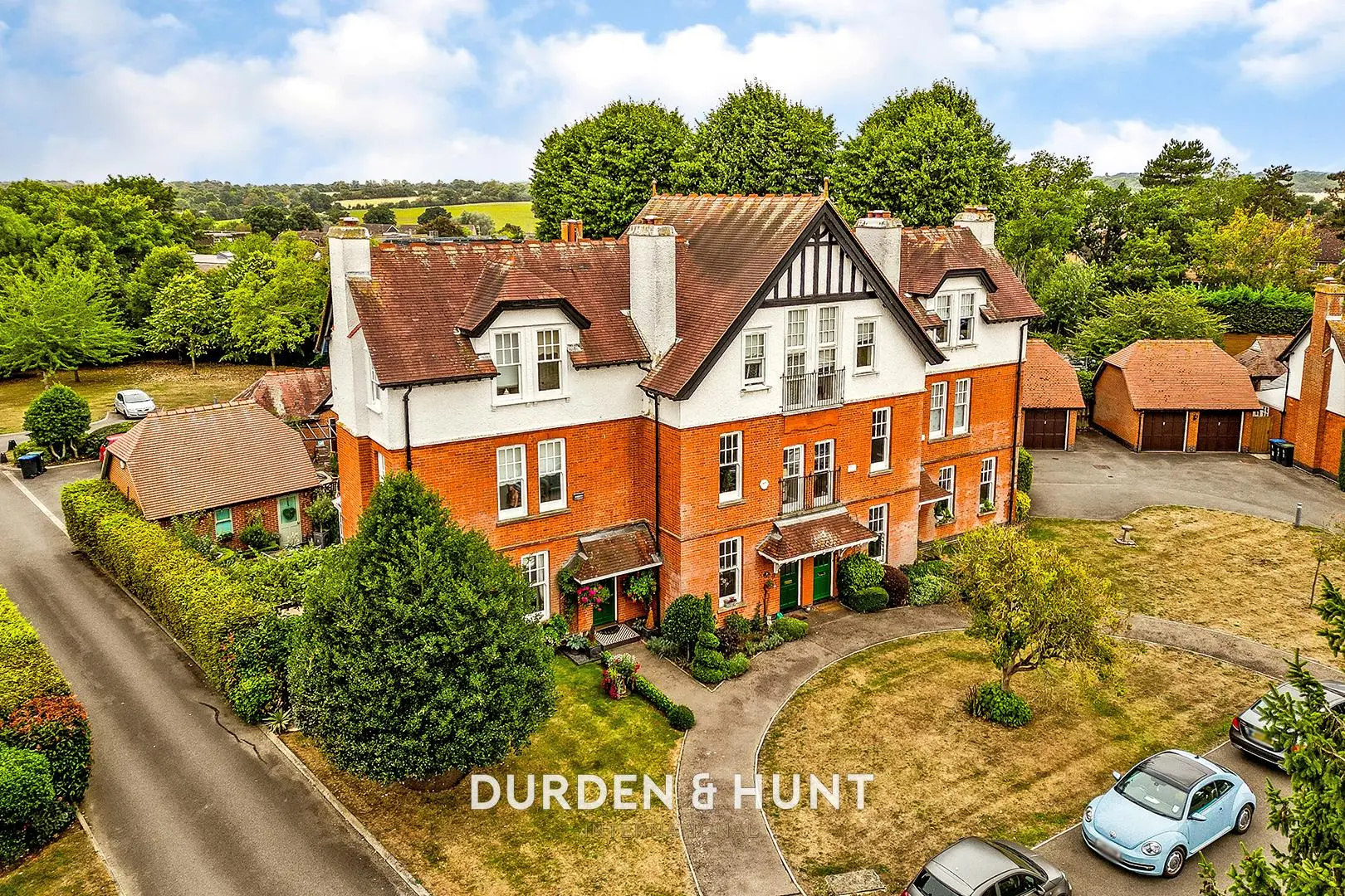 Aerial view of a large brick and white-rendered country house with red-tiled roof, surrounded by mature trees and manicured grounds, with parked cars visible. Durden & Hunt branding overlaid.