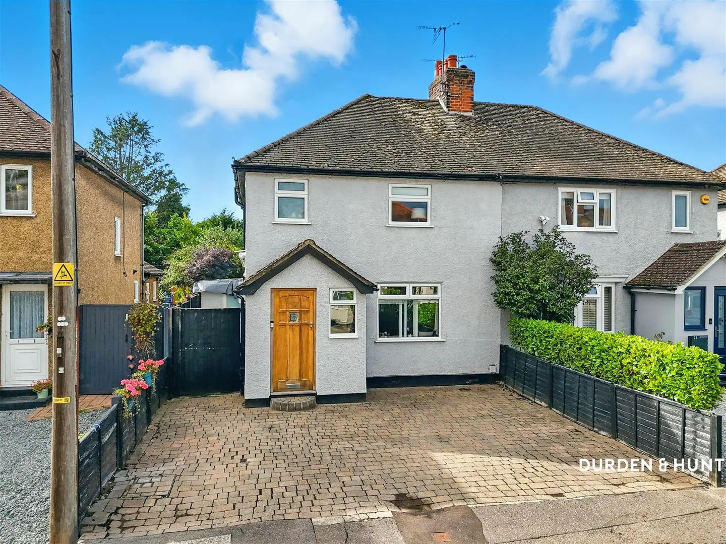 White semi-detached house with wooden front door, brick driveway, black fencing, and pitched roof with red chimney