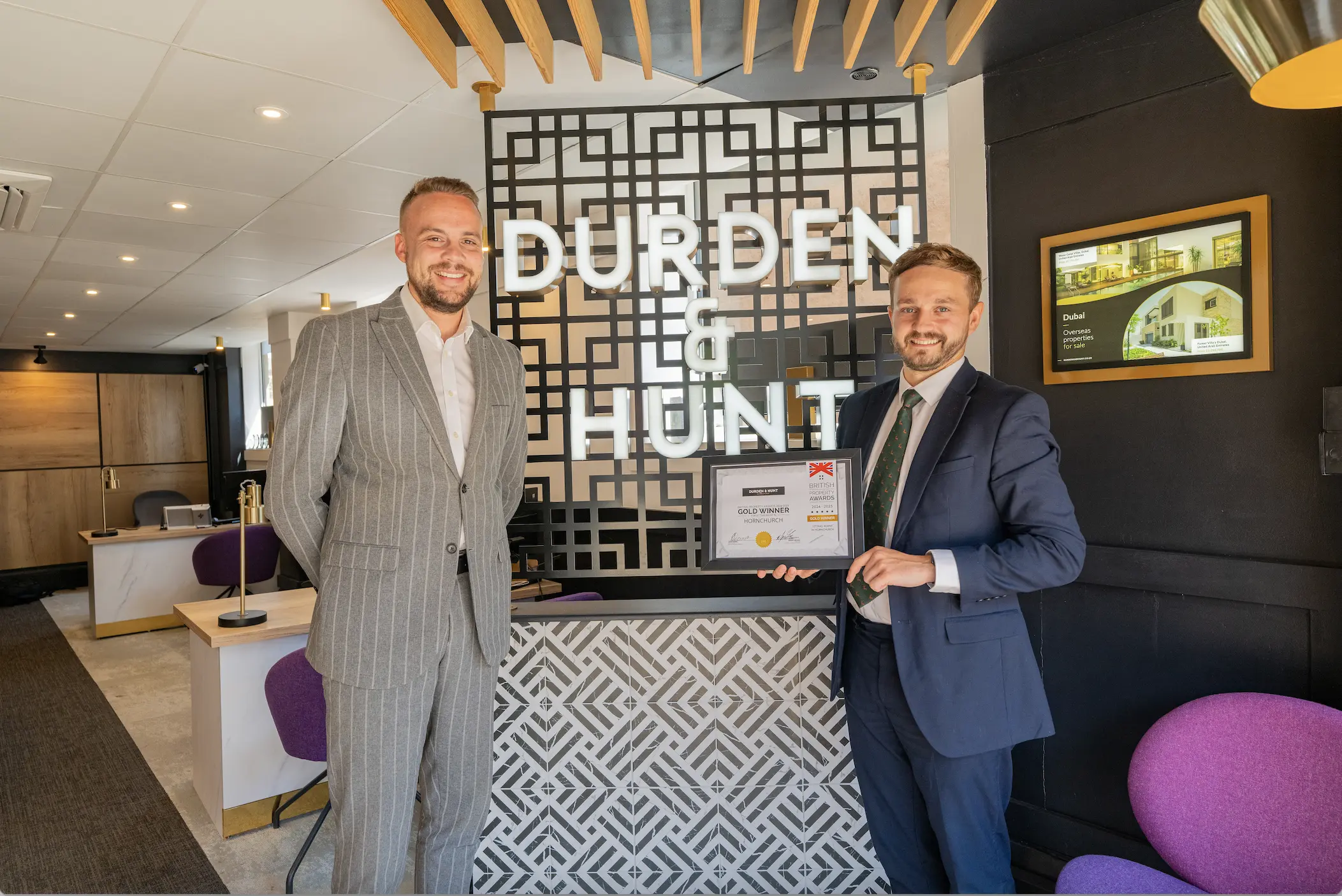Two men in business suits holding a certificate in a modern office with "DURDEN" signage and geometric wall patterns