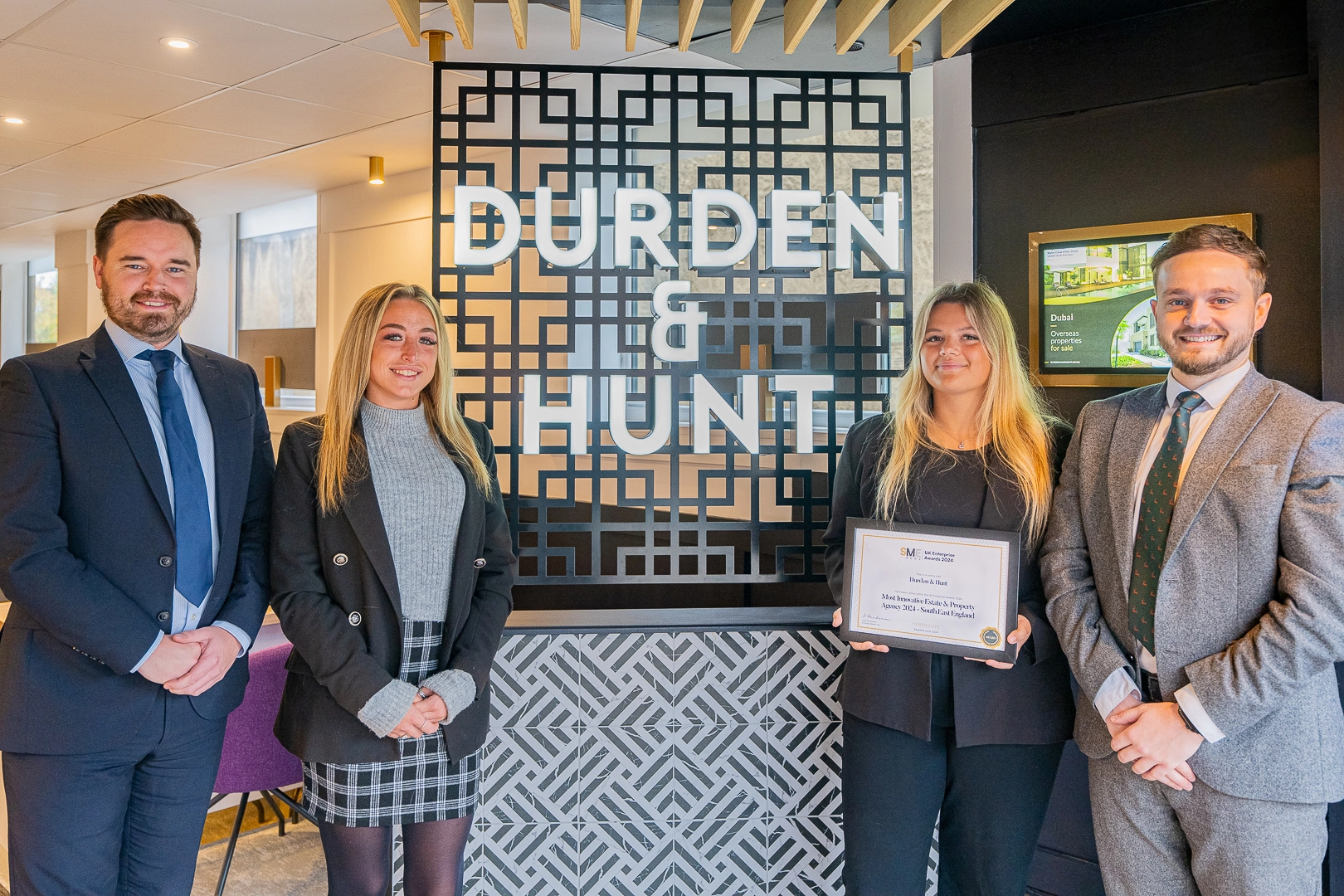 Four people in business attire standing in front of a Durden signage display, one holding a framed certificate