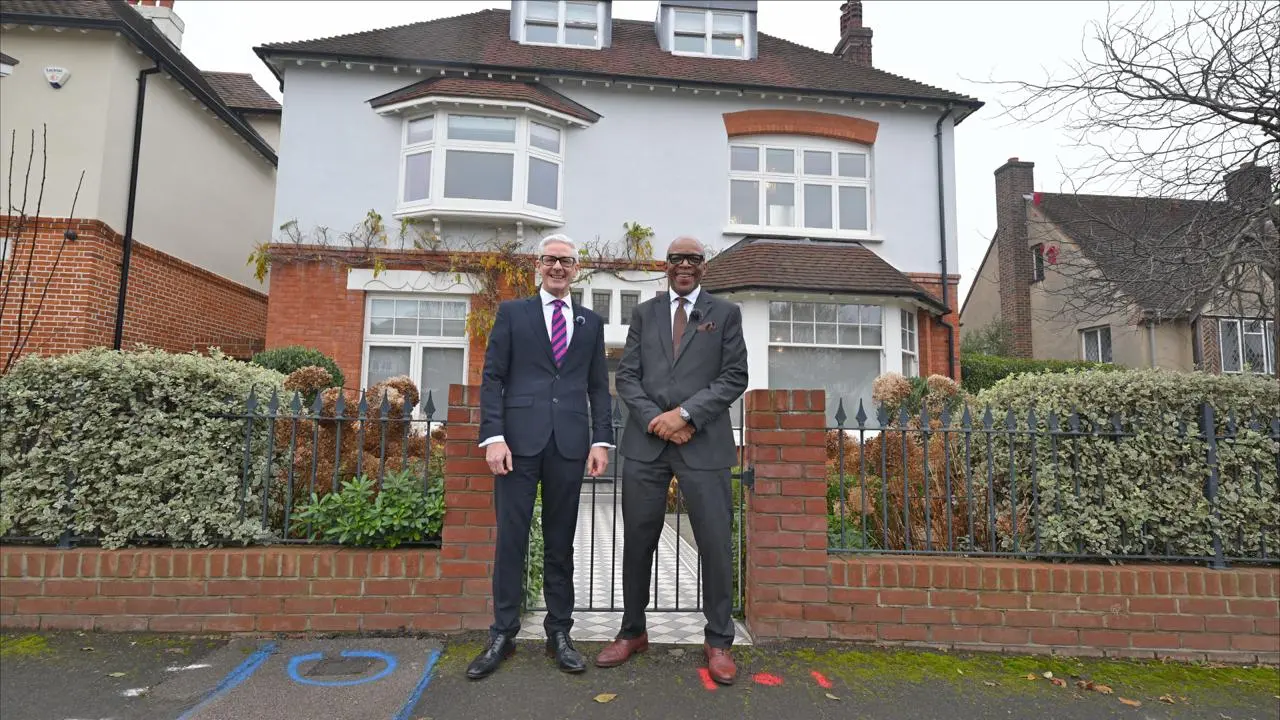 Two men in dark suits standing outside a white and red brick detached house with bay windows