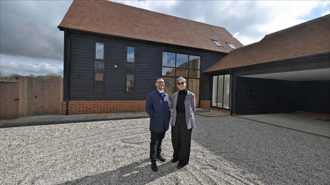 Two people standing in front of a modern black-clad house with brick base, large windows, and gravel driveway