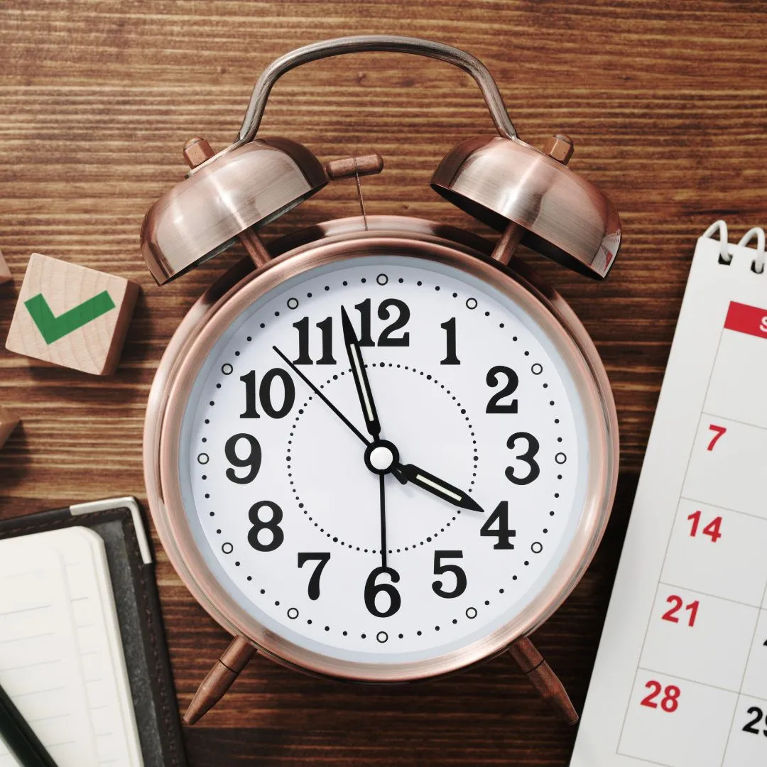 Copper twin-bell alarm clock showing 3 o'clock, surrounded by a calendar, notebook, and wooden desk