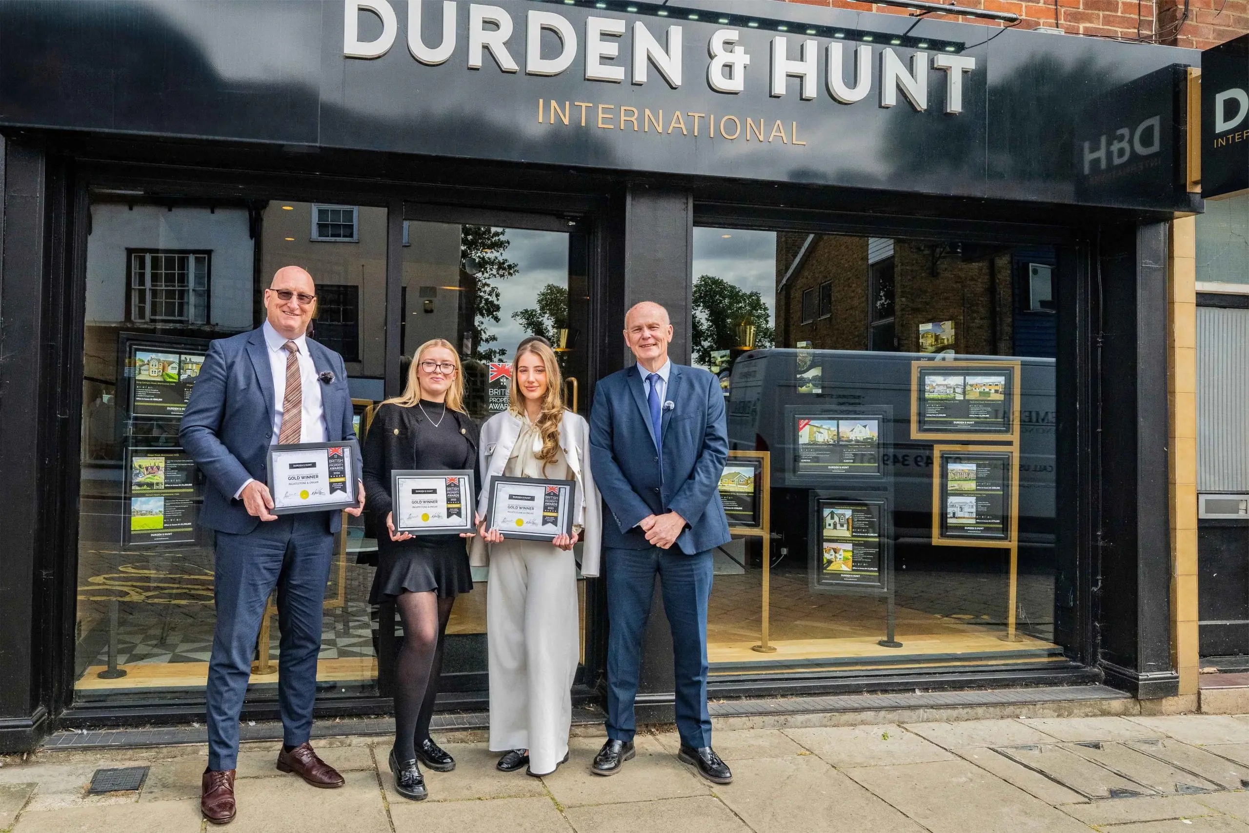 Four people holding framed certificates standing outside a Durden & Hunt International estate agency storefront