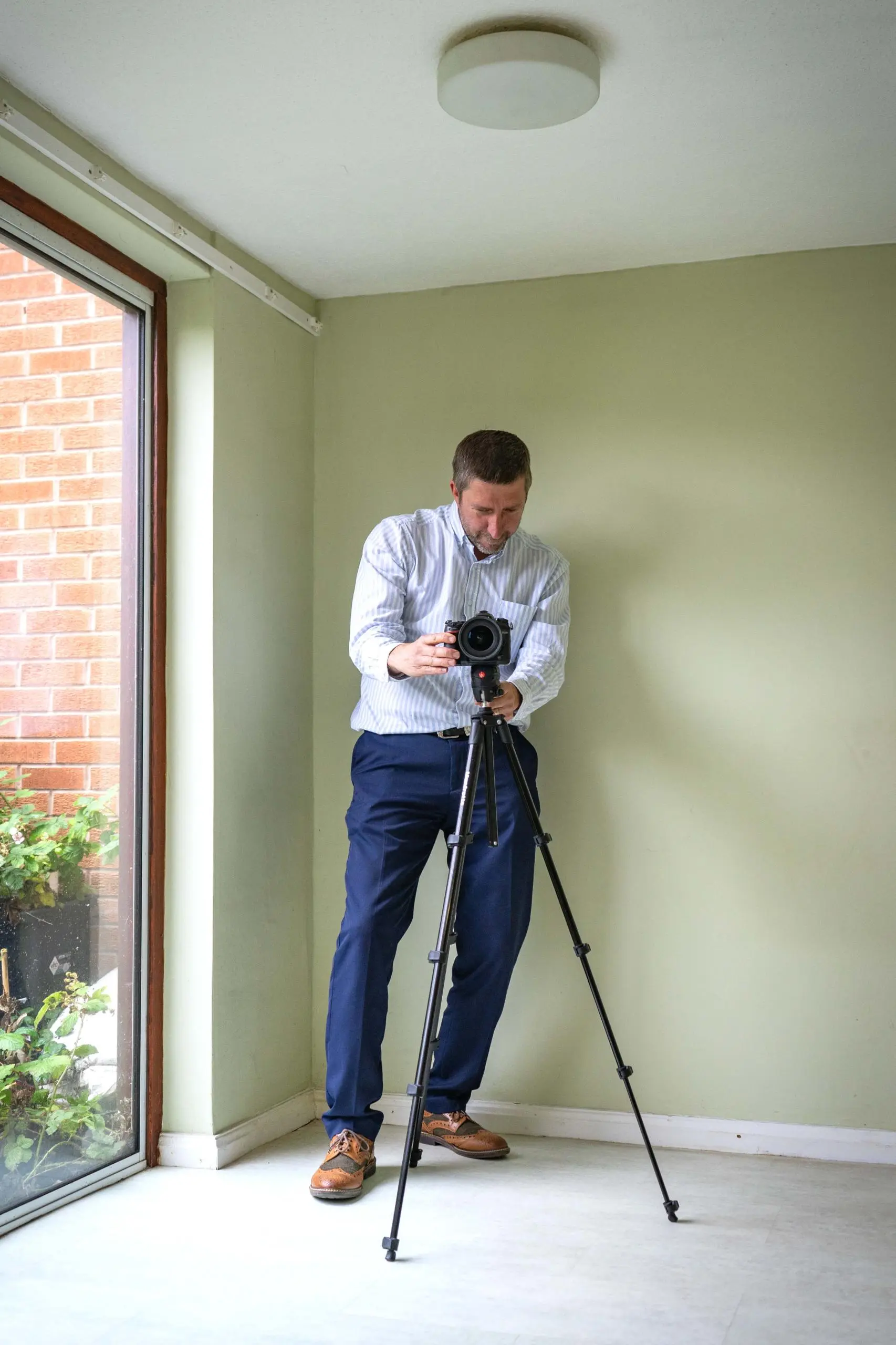Man with camera on tripod in pale green room with large window showing brick exterior