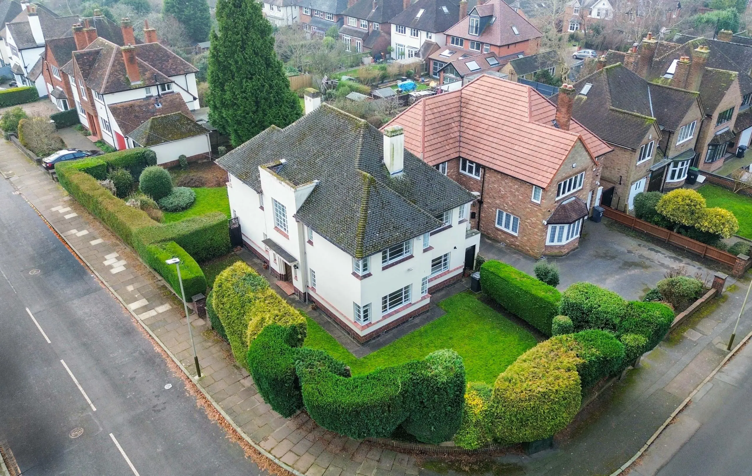 Aerial view of a white and red-brick residential property with dark roof and manicured hedges, surrounded by neighbouring houses on a tree-lined suburban street