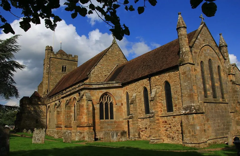 Stone church with tower, Gothic windows and pitched roof, surrounded by gravestones and green lawn under blue sky