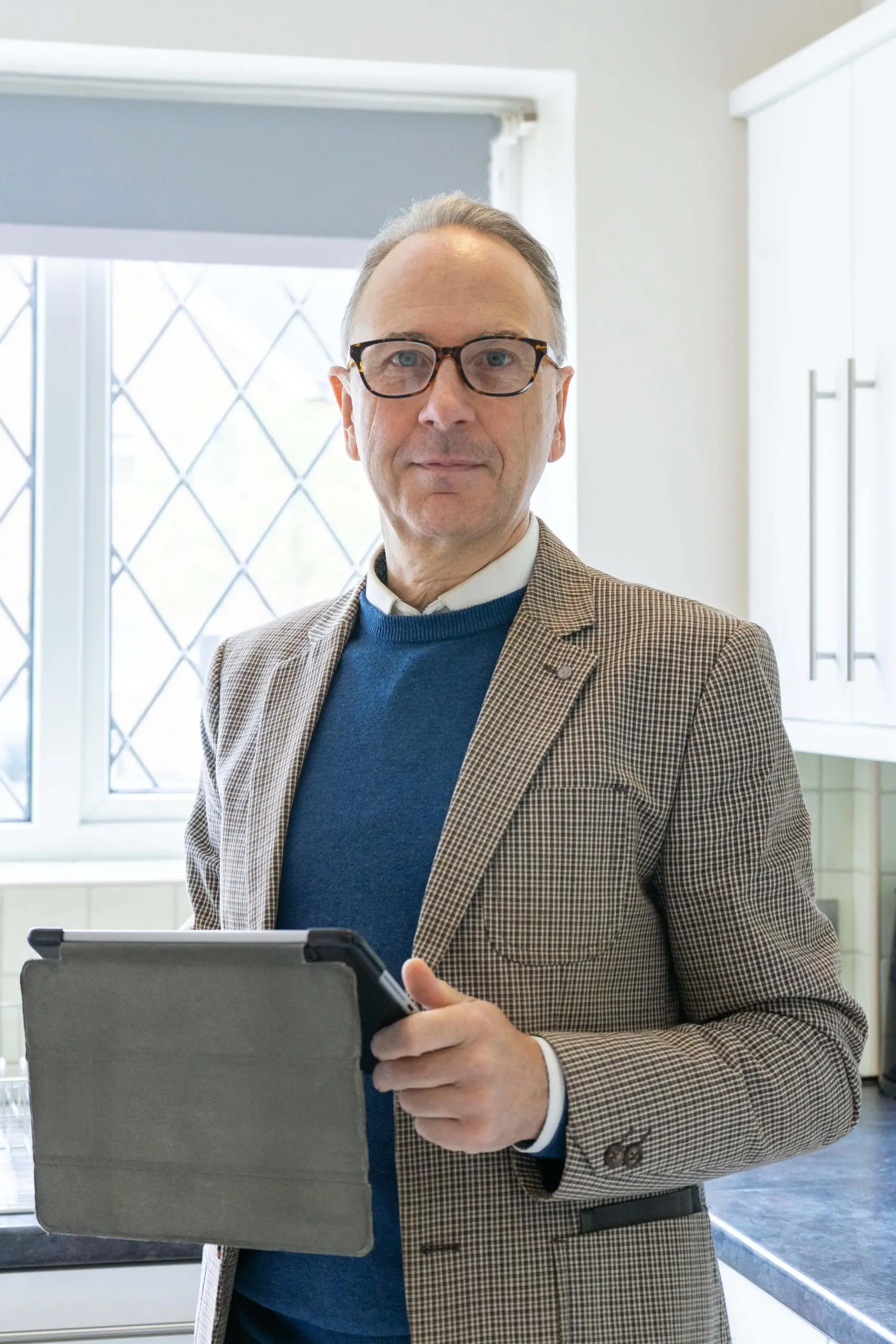 Man wearing glasses, beige checked blazer and blue jumper, holding a tablet, standing in a bright interior with white-framed windows