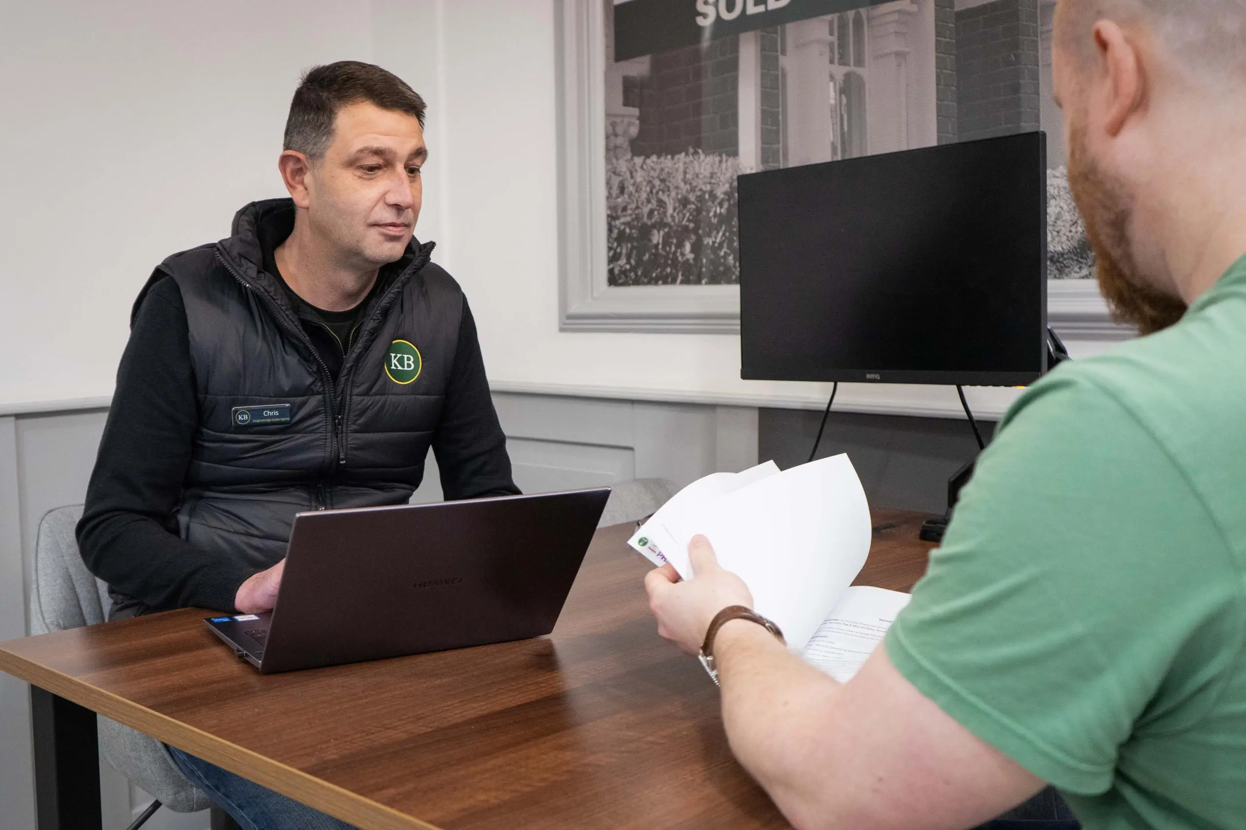 Two people at a desk with a laptop and papers; one wears a black KB-branded gilet, the other wears a green shirt